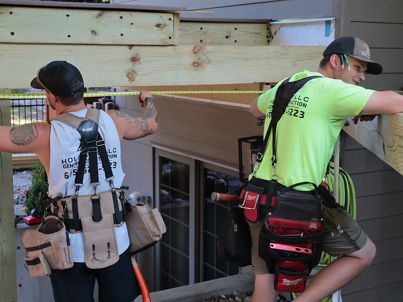 Two Hols & Co crew members in tool rigs at the back of a house, one running a tape along a new ledger board, both in shirts printed with the Hols & Co. LLC General Construction logo
