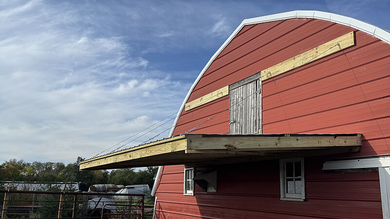 The gable end of a red steel-sided barn with an arched roof: a new pressure-treated lumber canopy with a metal roof, hung on steel cables from boards fixed across the gable on either side of the loft door, with a pipe livestock gate at the lower left