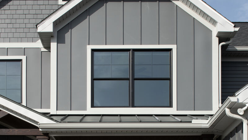 Gray board-and-batten siding on a gable with gray shingle siding on the wall behind it, wide white trim around a black-framed double window, white fascia, gutters and downspouts, and a dark standing-seam metal roof below, photographed by AZEK.