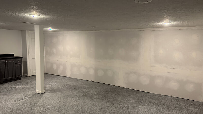Basement room mid-build with new gray carpet, a white-wrapped support post, recessed ceiling lights, a long drywall wall with mudded seams not yet painted, and a dark wet-bar cabinet run with a black countertop