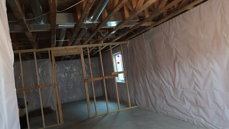 A below-grade room with new stud walls framed in front of foundation walls covered in plastic sheeting, open-web floor trusses and metal ductwork running overhead, a small window framed into the wall, and a bare concrete floor with a coiled green hose.