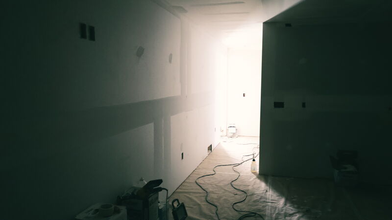 Basement hallway at the drywall stage, dark in the foreground, with light pouring in from a room at the far end across the taped seams and the paper-covered floor, where extension cords snake toward a work light