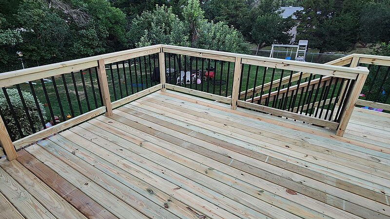 New pressure-treated deck boards running the length of a finished deck at dusk, with black metal balusters on the railing and a back yard beyond