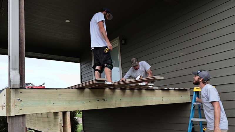 Three crew members on a new covered deck at a gray lap-sided house: one standing on a stack of composite boards, one crouched setting boards over the joists, and one on a blue stepladder at the new rim beam, with a downspout and the neighborhood beyond.