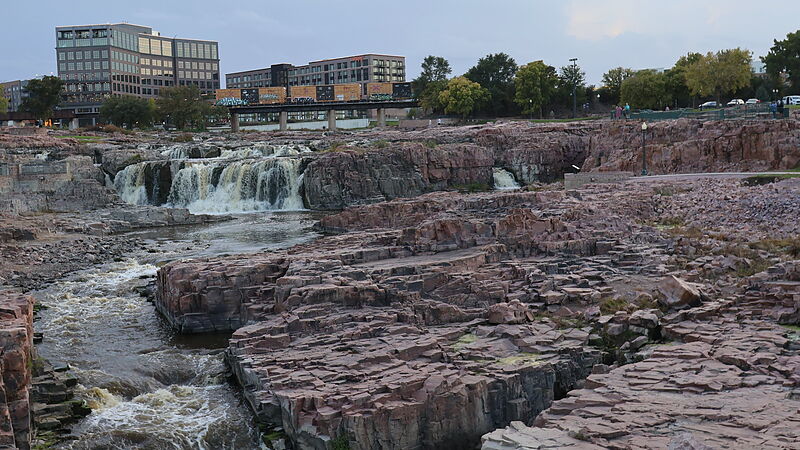 The falls of the Big Sioux River in Falls Park, Sioux Falls: white water pouring over ledges of pink quartzite, with downtown buildings and a freight train crossing a rail bridge behind, under a cloudy sky