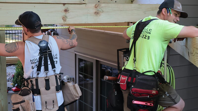 Two Hols & Co carpenters in branded shirts running a tape measure along a new header fastened to the house wall