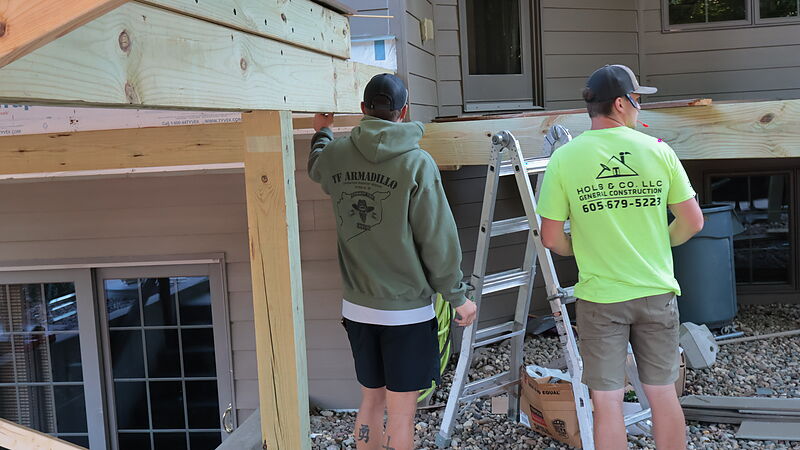 Two workers seen from behind at the side of a house, one reaching up to hold a wood beam against the wall above a post while the other stands at a folding ladder; the man at the ladder wears a lime-green shirt printed with the Hols & Co. LLC General Construction logo and a phone number.