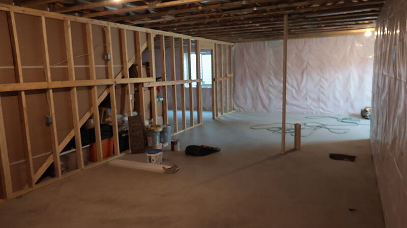 A framed lower level: a long stud partition wall with an opening on the left, foundation walls wrapped in plastic sheeting on the right, open joists and metal ductwork overhead, and a bare concrete floor with a coiled hose.