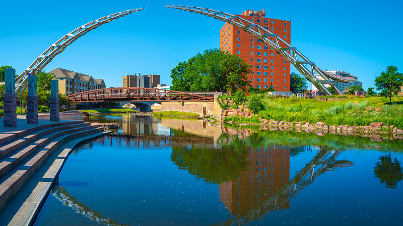 A curved stainless-steel arch sculpture spanning a calm blue river in downtown Sioux Falls, with a rust-red truss footbridge, a tall red-brick tower and granite riverside steps reflected in the water under a clear summer sky