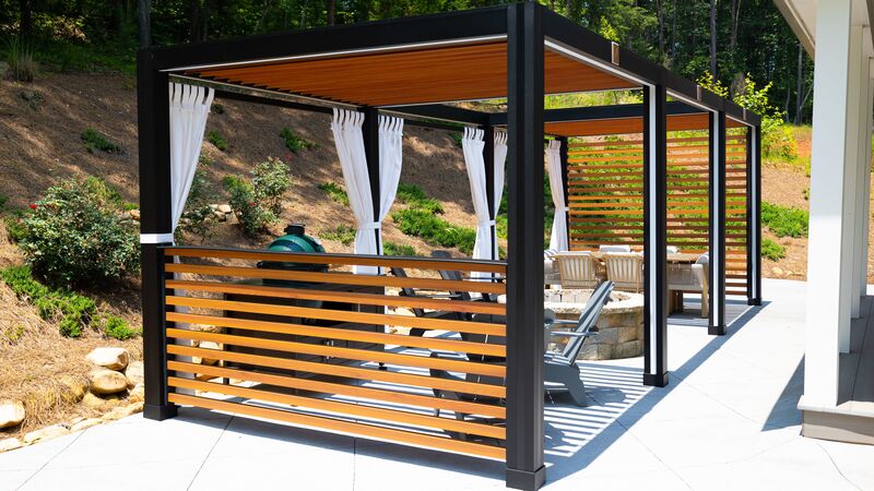 A black-framed pergola with wood-look louvers overhead and wood-look privacy slats on two sides, white curtains tied back at the posts, a green kamado grill behind the slats, gray Adirondack chairs at a round stone fire pit and a dining set beyond, on a concrete patio below a wooded hillside, photographed by StruXure.
