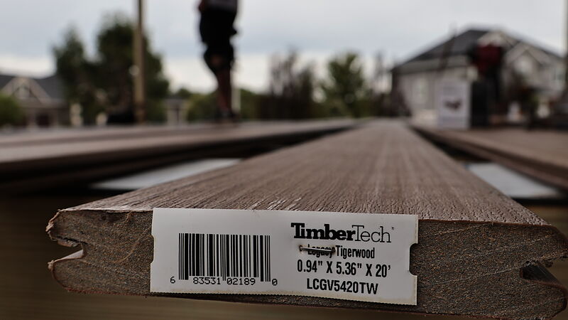 Close-up of the end of a TimberTech Legacy Tigerwood composite deck board with its product label still on, laid across the joists of a deck under construction, a crew member's legs and a gray two-story house out of focus beyond