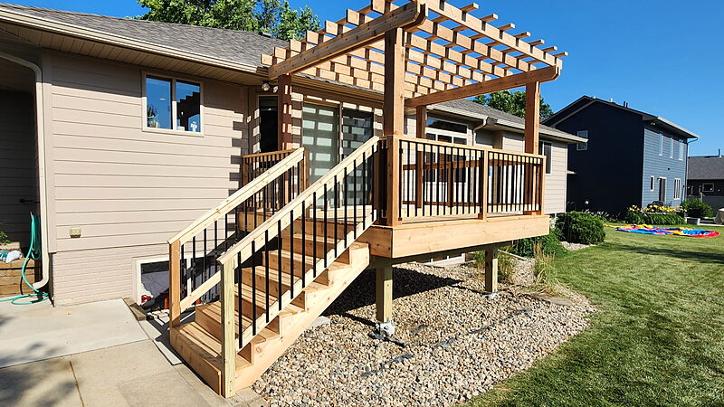 Finished pressure-treated deck with a lattice-top pergola and black metal balusters, a stair run down to a concrete pad, and support posts set in a rock bed