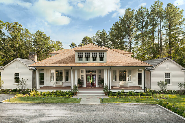 A white clapboard house with a wide front porch decked in warm brown boards, white columns, lanterns and a wood front door, a cedar-shingle roof with a dormer, and a gravel drive and lawn in front, photographed by TimberTech.