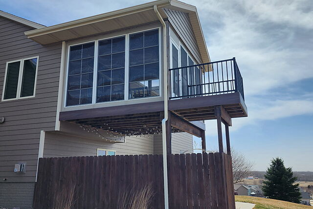 Second-story sunroom addition with an aluminum-railed balcony deck, built over a fenced patio strung with lights