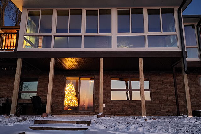 Straight-on dusk view of a second-story sunroom addition over a stone walkout level, a lit Christmas tree visible through the patio door