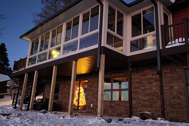 Second-story sunroom addition with window walls lit from inside at dusk, carried on posts over a stone-clad walkout level with snow on the ground