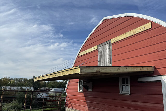The gable end of a red steel-sided barn with an arched roof: a new pressure-treated lumber canopy with a metal roof, hung on steel cables from boards fixed across the gable on either side of the loft door, with a pipe livestock gate at the lower left