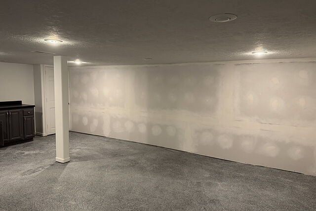 Basement room with new gray carpet, a white-wrapped support post, recessed ceiling lights, a long drywall wall with mudded seams not yet painted, a dark wet-bar cabinet run with a black countertop, and a white panel door