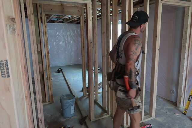 A crew member in a cap, sleeveless shirt and tool belt standing among freshly framed stud partitions in a lower level, a bucket and a level on the concrete floor, plastic-wrapped foundation wall and open joists beyond.