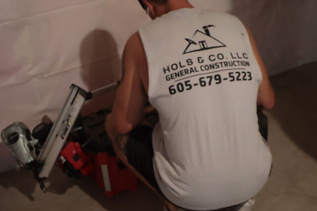 A crew member in a white Hols & Co. LLC General Construction shirt crouched at a circular saw and a red tool case on the bare concrete floor, plastic-wrapped foundation wall behind.