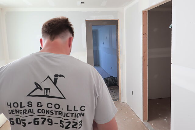 A crew member in a gray Hols & Co. LLC General Construction shirt seen from behind, walking through a drywalled lower level with two cased doorways ahead and tools on the floor.