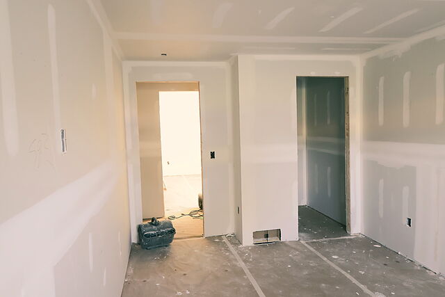 A drywalled lower-level hallway with two cased door openings into the next rooms, seams taped and screw heads mudded, a shop vacuum inside one doorway and the floor still bare.