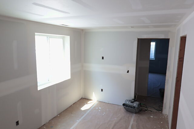 The same lower-level room three weeks on: drywall hung and taped on every wall, the window at left, a doorway through to a second room, a dehumidifier on the paper-covered floor.