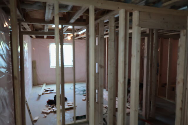 A framed lower-level room seen through its stud partition walls: a window in the far wall, foundation walls wrapped in plastic, cut lumber and offcuts on the bare concrete floor, open joists and ductwork overhead.