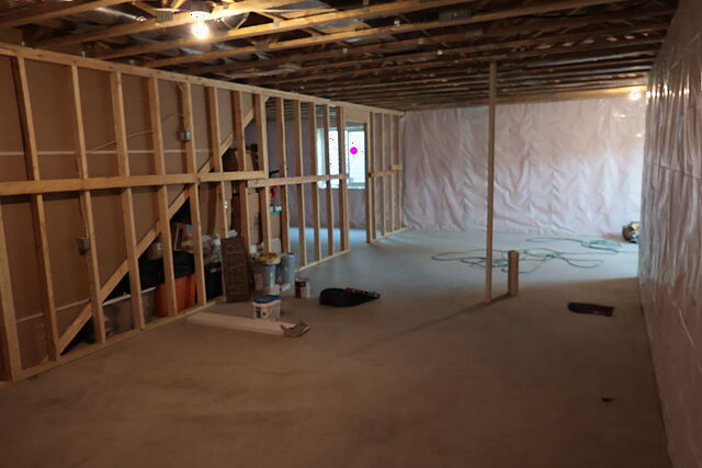 A long view down a framed lower level: a run of stud wall on the left with openings framed into it and storage bins tucked in the open bays, a window in the framing at the far end, foundation walls wrapped in plastic on the right, and buckets, a tool bag and a coiled hose on the bare concrete floor.