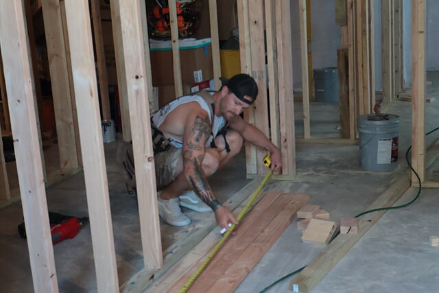A worker in a tool belt and backwards cap crouched on a bare concrete floor, pulling a tape measure along boards laid out beside a framed stud wall, with scrap blocks, a five-gallon bucket and a tool bag nearby.