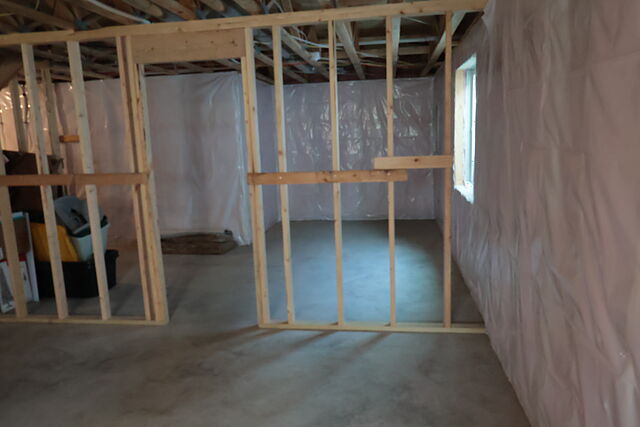 Stud partition walls framed on a bare concrete floor in a lower level, a doorway opening framed through one wall, foundation walls covered in plastic sheeting, open joists overhead, and a window at the far right.