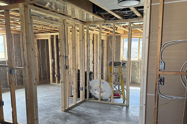 Unfinished basement with new wood stud walls on a bare concrete floor, electrical boxes and white cable run through the studs, under an open joist ceiling with recessed light cans