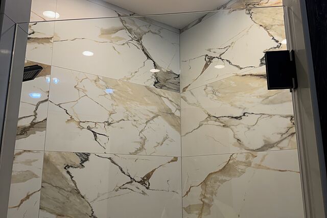 Looking up inside a shower finished in large-format marble-look tile carried to the ceiling, with a black square rain head, black handle, and two recessed ceiling lights