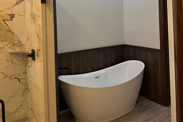 Freestanding white slipper tub in a wood-paneled alcove beside a marble-look tile shower with a black rain shower head and a glass panel