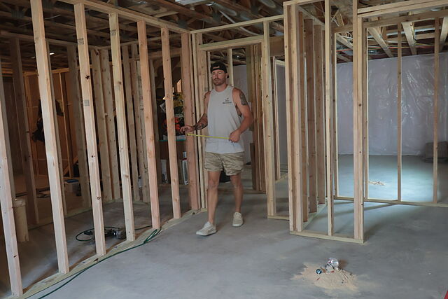 A man in a white sleeveless shirt printed with the Hols & Co. LLC logo pulling a tape measure across a framed opening in a below-grade level, with stud partition walls, a bare concrete floor, plastic sheeting on the far wall and open joists overhead.