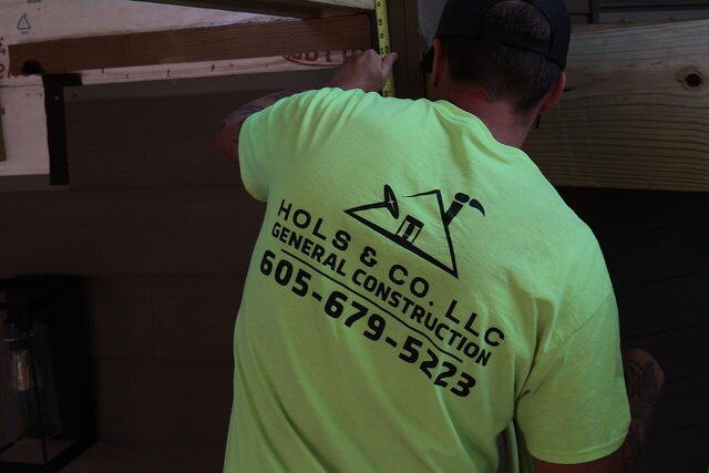 Hols & Co crew member in a company shirt printed HOLS & CO. LLC GENERAL CONSTRUCTION 605-679-5223, running a tape along a header beam