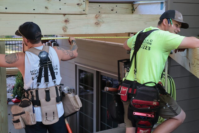Two Hols & Co carpenters in branded shirts running a tape measure along a new header fastened to the house wall