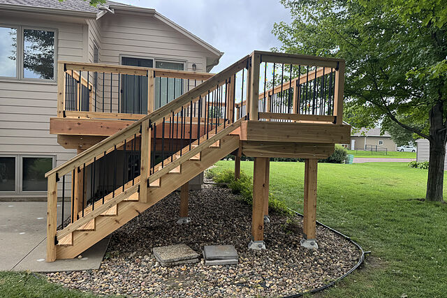 Side view of an elevated cedar-toned deck with black-baluster rails on a beige lap-sided split-level house, a straight stair down to a concrete patio, posts set in a river-rock bed