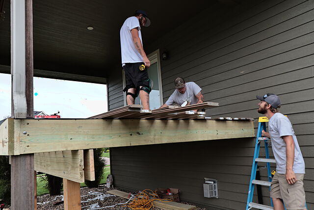Three workers on and around a new elevated deck against the back of a house. One stands on the frame near the door, a second kneels at the edge setting brown grooved deck boards over the joists, and a third stands on a blue stepladder alongside the new rim joist.