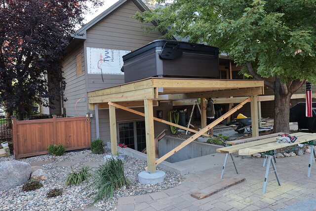 Elevated deck frame on 6x6 posts with temporary bracing, a hot tub already set on the framing, and a Tyvek-wrapped wall behind