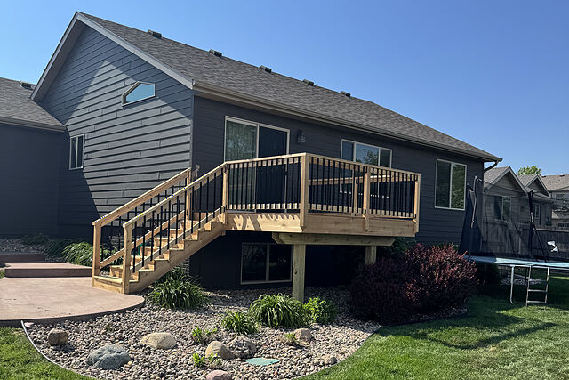Charcoal lap-sided house with a new unstained wood deck and black metal balusters, a straight stair down to a colored concrete patio, and deck posts standing in a river-rock bed with shrubs