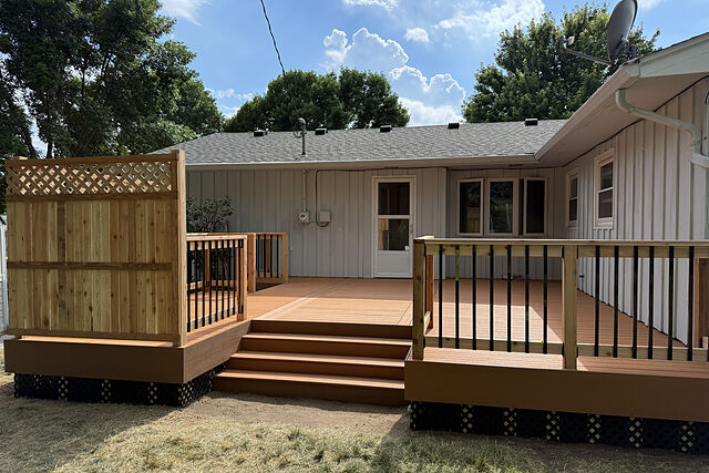 Low two-level composite deck in a brown-orange tone across the back of a white board-and-batten ranch house, with wide center steps, black metal balusters in wood rails, and a cedar privacy screen topped with lattice at one end