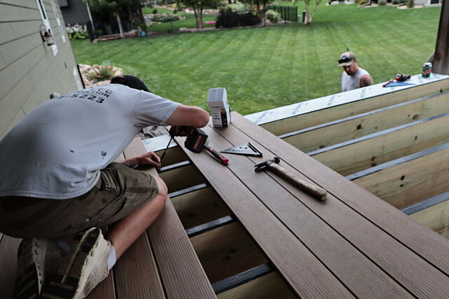 A worker in a Hols & Co shirt kneeling on new brown composite deck boards, driving a fastener with a cordless drill, a box of fasteners, a speed square and a hammer beside him, open joists ahead of him, and two more crew at the far edge of the deck over a mown lawn.