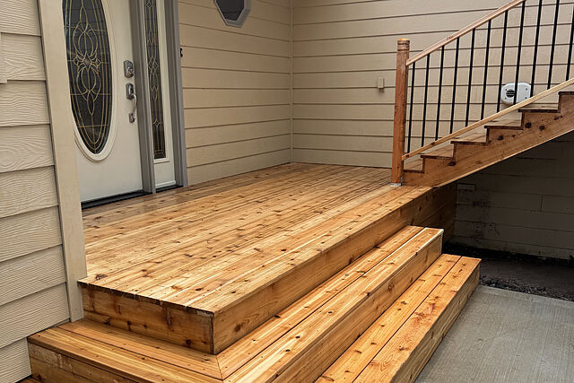 Cedar-toned wood landing at a front door with an oval leaded-glass insert, three wraparound box steps down to a concrete driveway, and a wood stair with black balusters climbing to the right against tan lap siding