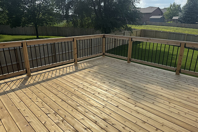 Deck surface of pale cedar-toned boards with wood rails and black metal balusters casting shadows, looking out over a fenced back yard with mature trees