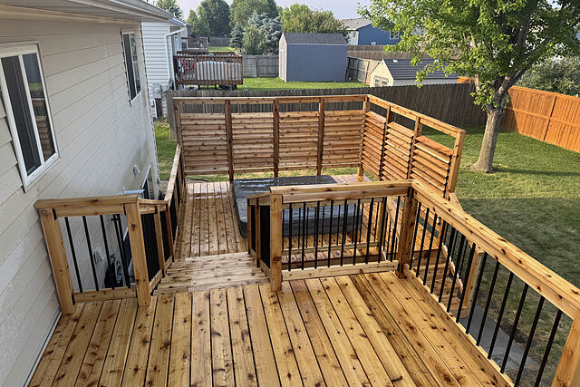 View from above an upper cedar deck with black metal balusters: a stair down to a lower deck where a covered hot tub sits behind louvered cedar privacy screens, fenced lawn and neighboring yards beyond