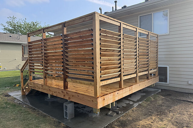 Low cedar deck on a concrete pad beside a tan lap-sided house, with tall louvered cedar privacy screens on two sides and a short stair up to a higher deck with black metal balusters