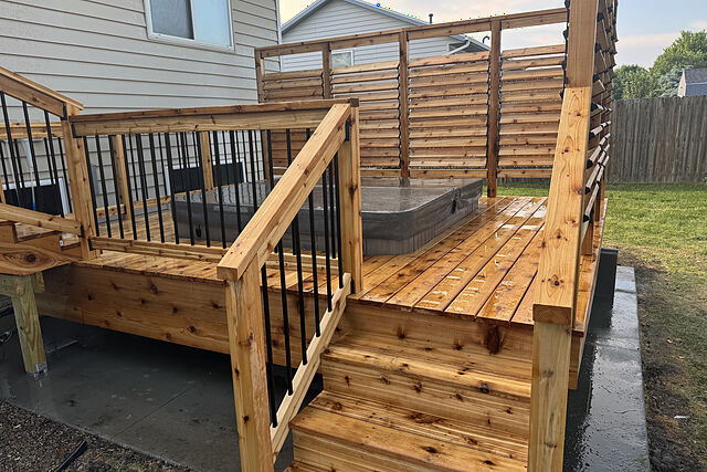 Cedar stair leading up onto a low deck where a covered hot tub sits behind black-baluster railings and louvered cedar privacy screens, beside a tan lap-sided house