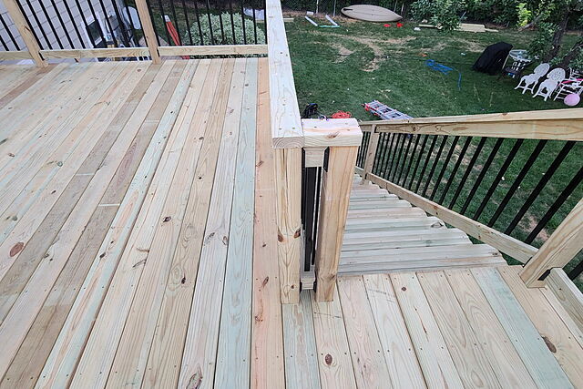 New pressure-treated deck boards and black-baluster railing at a corner stair transition, ladders and tools still in the yard below