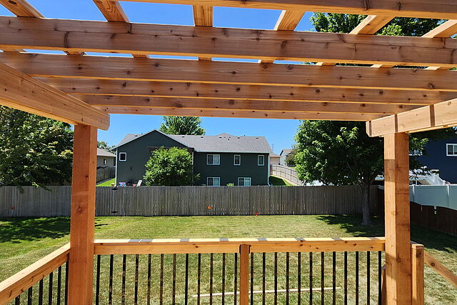 Cedar pergola rafters over a deck railing with black metal balusters, looking out across a fenced back yard